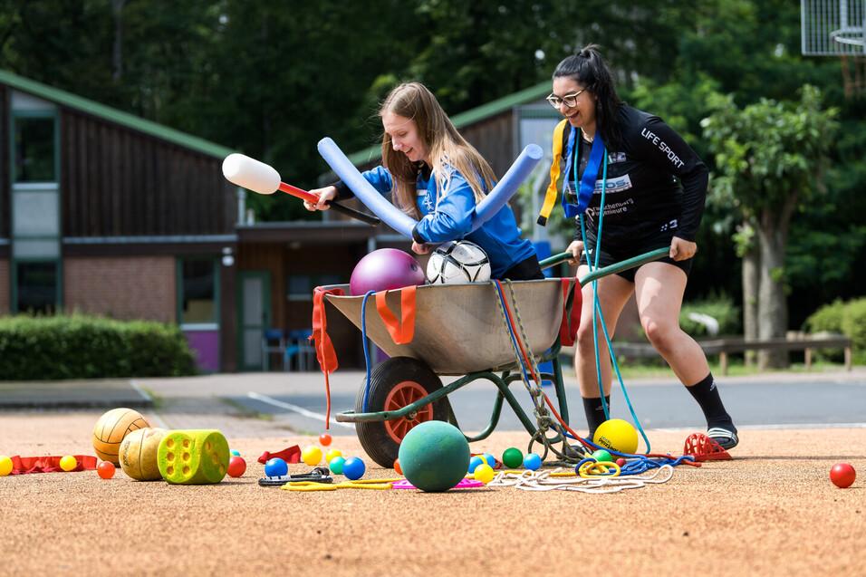 Mädchen schiebt Mädchen im Schubkarre mit vielen Spielutensilien