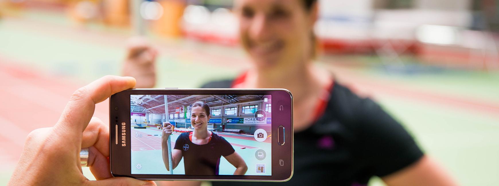 Hand hält ein Smartphone, das ein Porträt einer lächelnden Athletin mit Stab in einer Sporthalle zeigt.